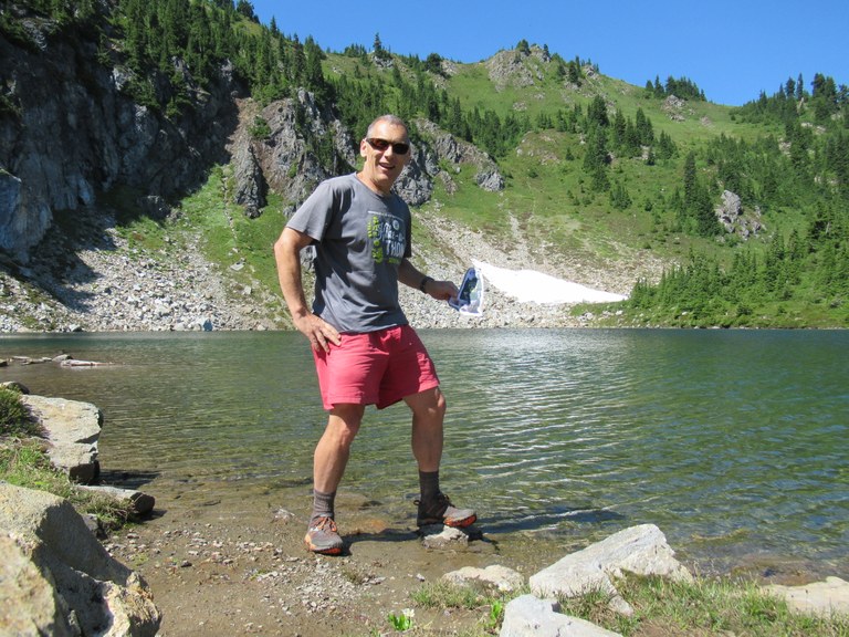 Craig poses for a photo with a cardboard Miles the Marmot. He stands on the rocky shore of an alpine lake.