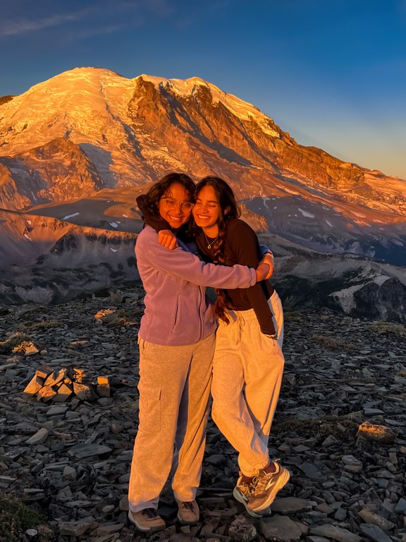 Two friends hug on rock landscape at sunset with snow capped Rainer in the background. 