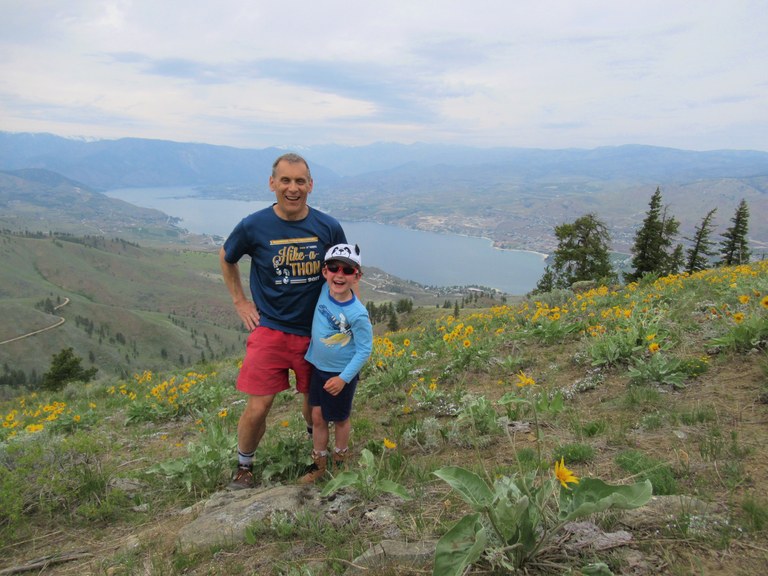 Craig and a child pose for a photo on a hillside dotted with wildflowers. Far below the hill spans a large body of water.