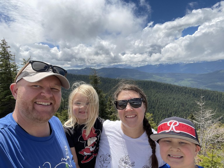 A family of four, two adults and two kids, take a selfie on an overlook above evergreen trees. 