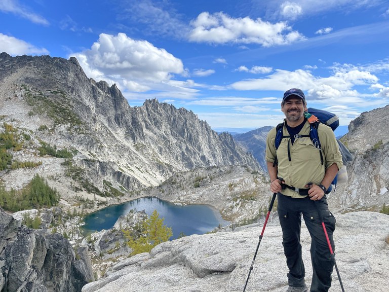 Jaime Loucky at the Enchantments by Cassidy Giampetro Jaime Loucky smiles from a rocky outcrop above an alpine lake in the Enchantments. He is wearing his backpack and has his trekking poles in his hands.