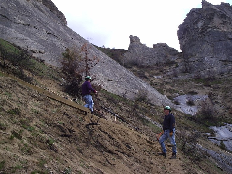 Two green hat trail volunteers haul equipment on a grass hillside.