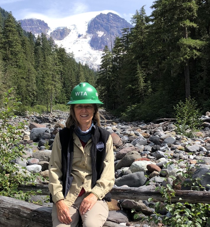 Kathy rests on trail near a big mountain. She's dressed in WTA volunteer garb with a big smile.