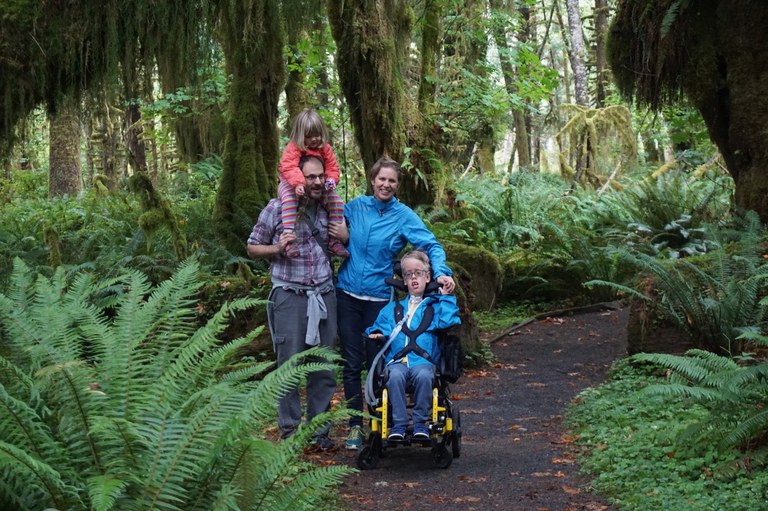 A family of hikers hike down a forested path. One child is in a wheelchair and is being pushed by a parent. The other child is on the other parent's shoulders.