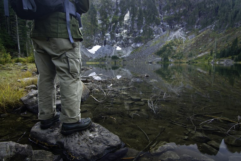 A hiker, shown from waist down, stands on a rock next to a mountain lake. 