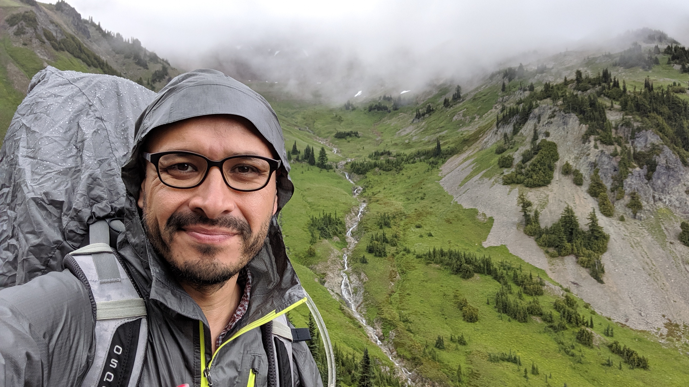Matt Martinez mountain selfie. Matt Martinez takes a selfie from a mountain pass. Mist engulfs the top of the mountains behind him and a waterfall tricks down its face. He is wearing a rain jacket and backpack.