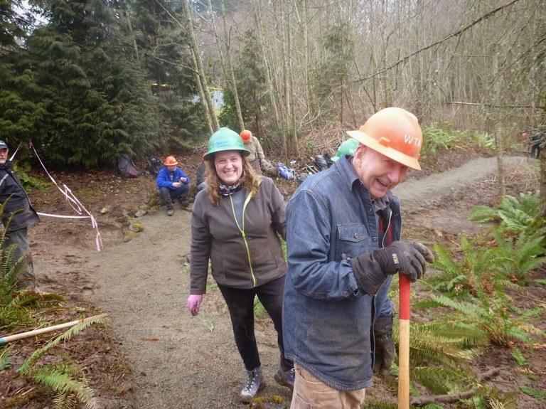 Pete smiles in the foreground of a photo while holding a shovel while a green hat behind him smiles. 