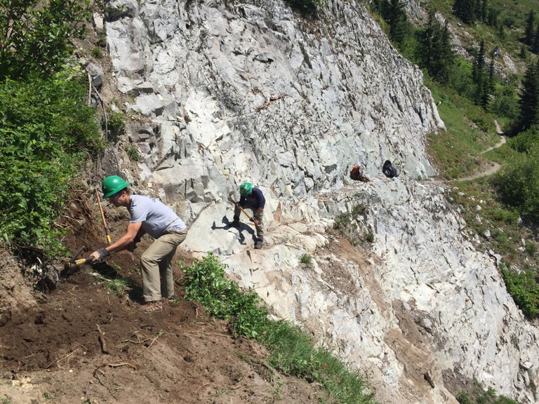 A group of WTA volunteers work on a trail traversing a steep side slope with lots of rock. 