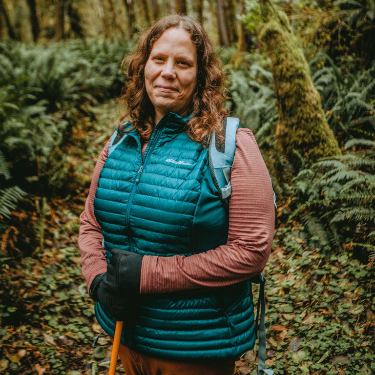 Syren on a forested trail wearing a bright puffy vest and holding a pair of trekking poles.