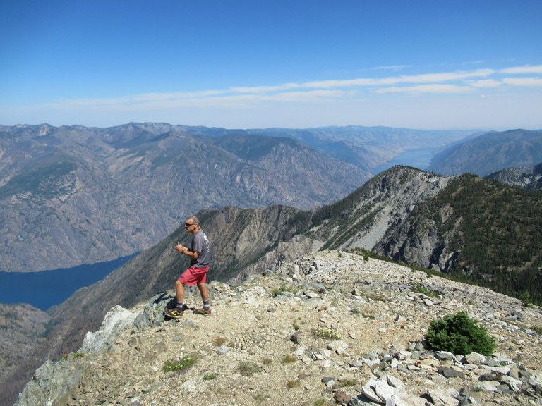Craig stands near a ledge on trail with a view down to a lake below.