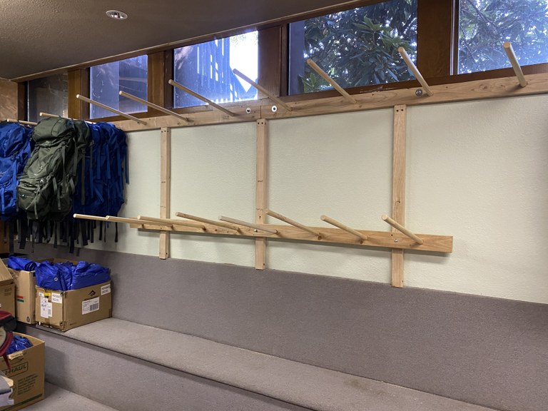 Wall of wooden pegs for backpack storage. Window above pegs. Green and blueovernight backpacks to the left of image.