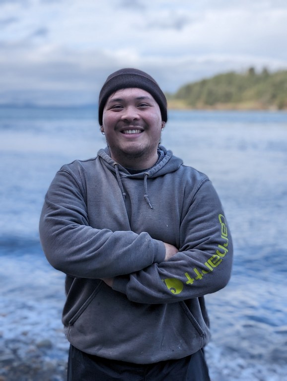 Riley poses with their arms crossed in front of the Salish Sea.