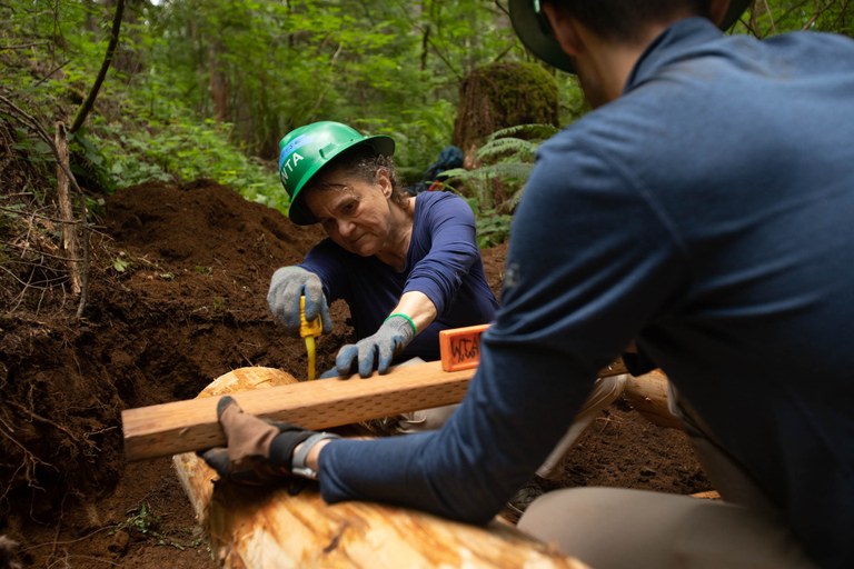 Two people work together on trail to measure wood wearing green hats.