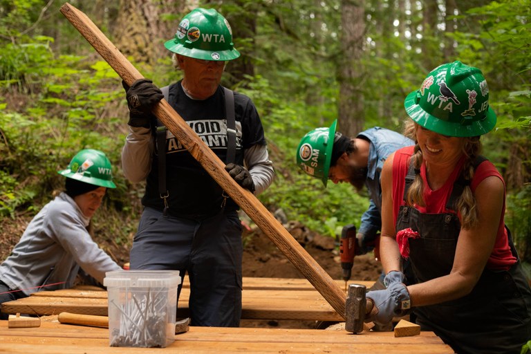 Four volunteers wearing green hats align pieces of wood while on trail.