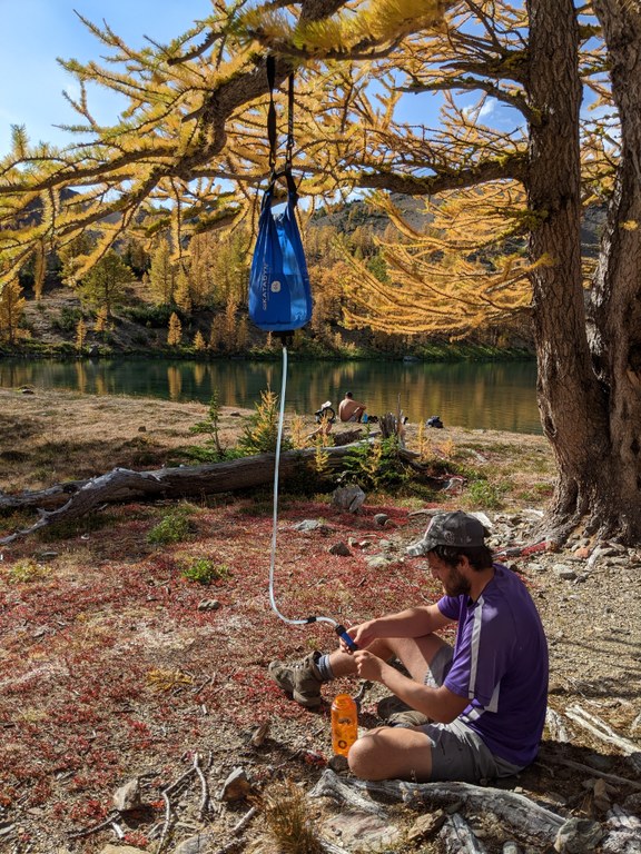 A hiker sites on the ground and connects their gravity filter to their water bottle.