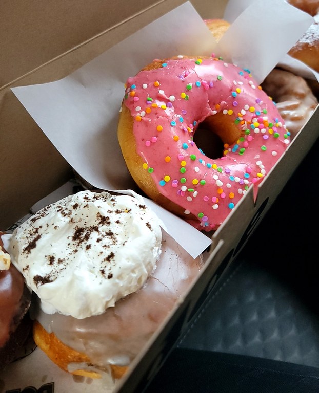 Two donuts are highlighted in a long, narrow box. One is lavender colored with whipped cream on top. The other is pink with sprinkles.