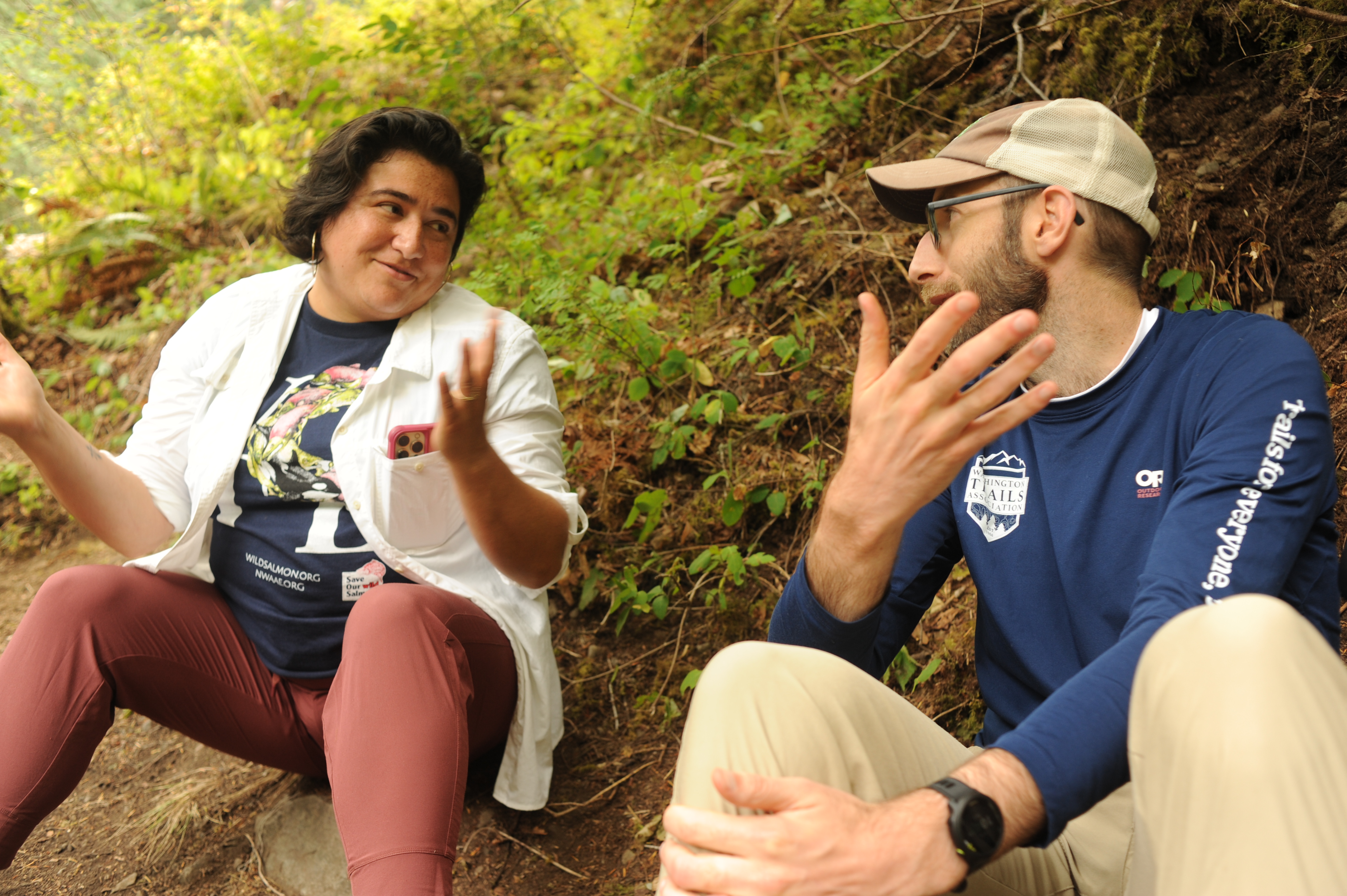 Two people, one in a Washington Trails Association shirt, sit and talk in the forest