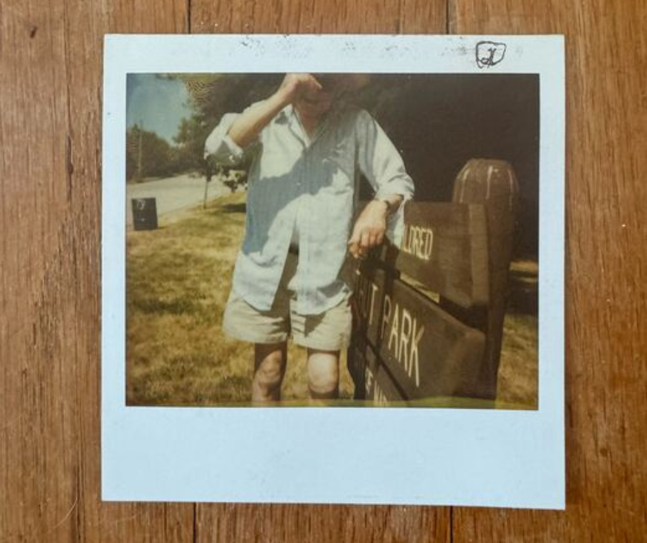 Photo of Polaroid on wood floor. Man pulls hat over head standing in front of frame.