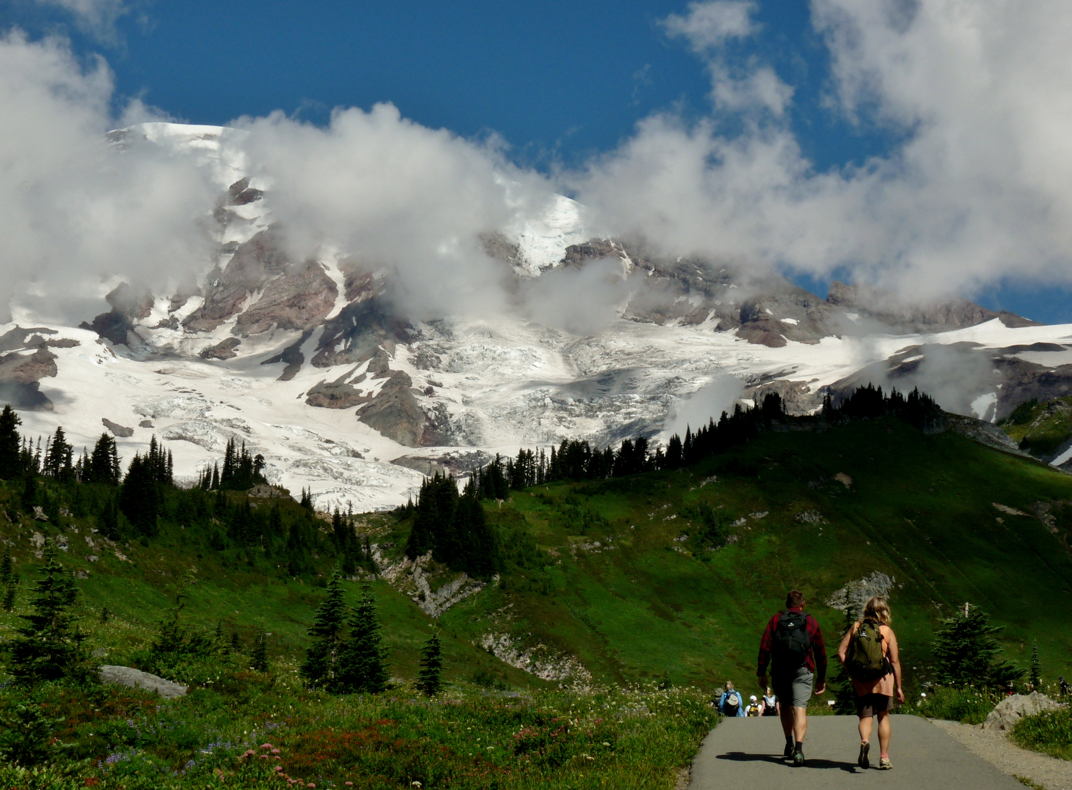 Two hikers seen from the back on a trail with Mount Rainier in the background