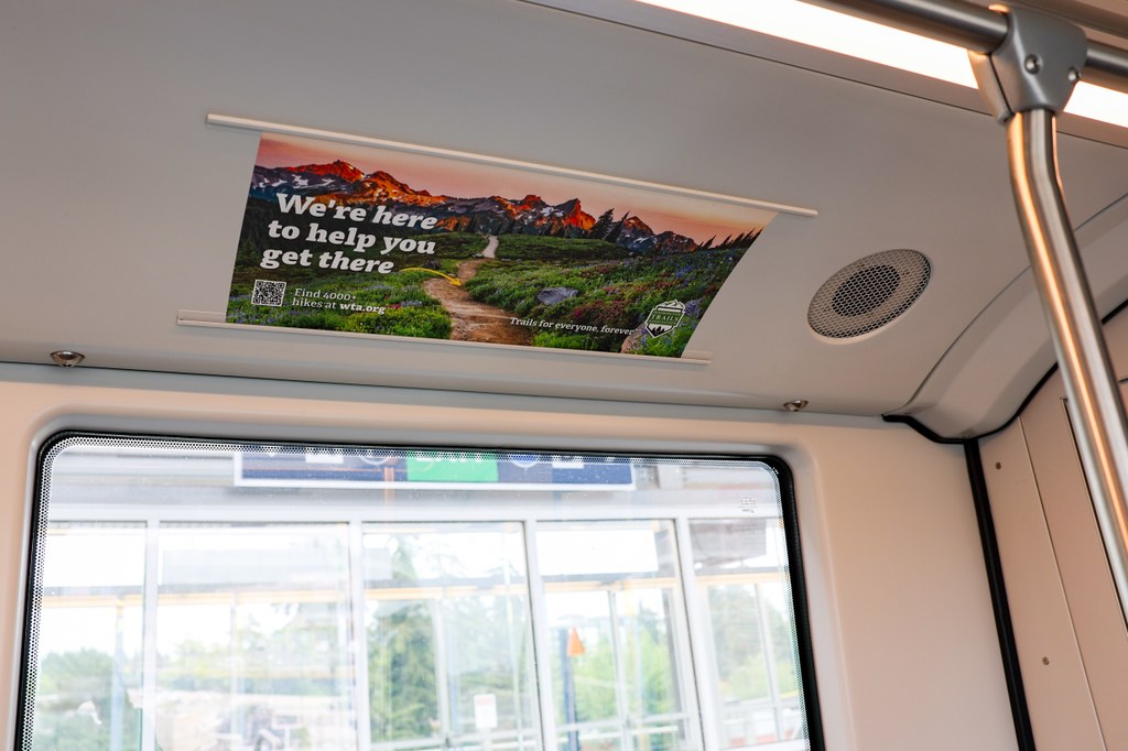WTA ad on Sound Transit. WTA advertisement on Link Light Rail. A wildflower-lined path at sunset along with text “We’re here to help you get there” with an arrow pointing to the trail.