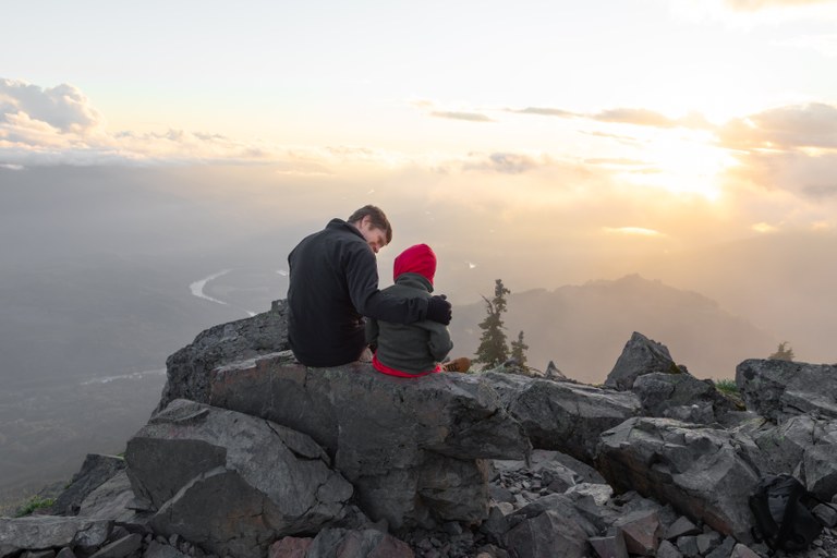 Two hikers sit on a rock, looking out over a mountainous landscape. 