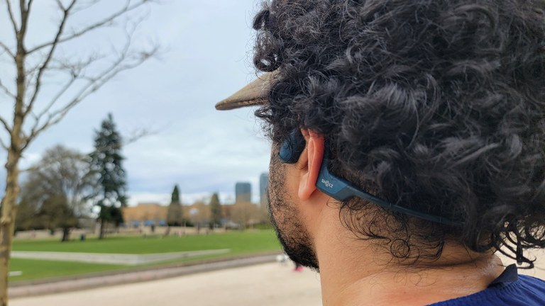 A closeup of a hiker wearing bone-conduction headphones under a visor and dark curly hair, looking out over a city park