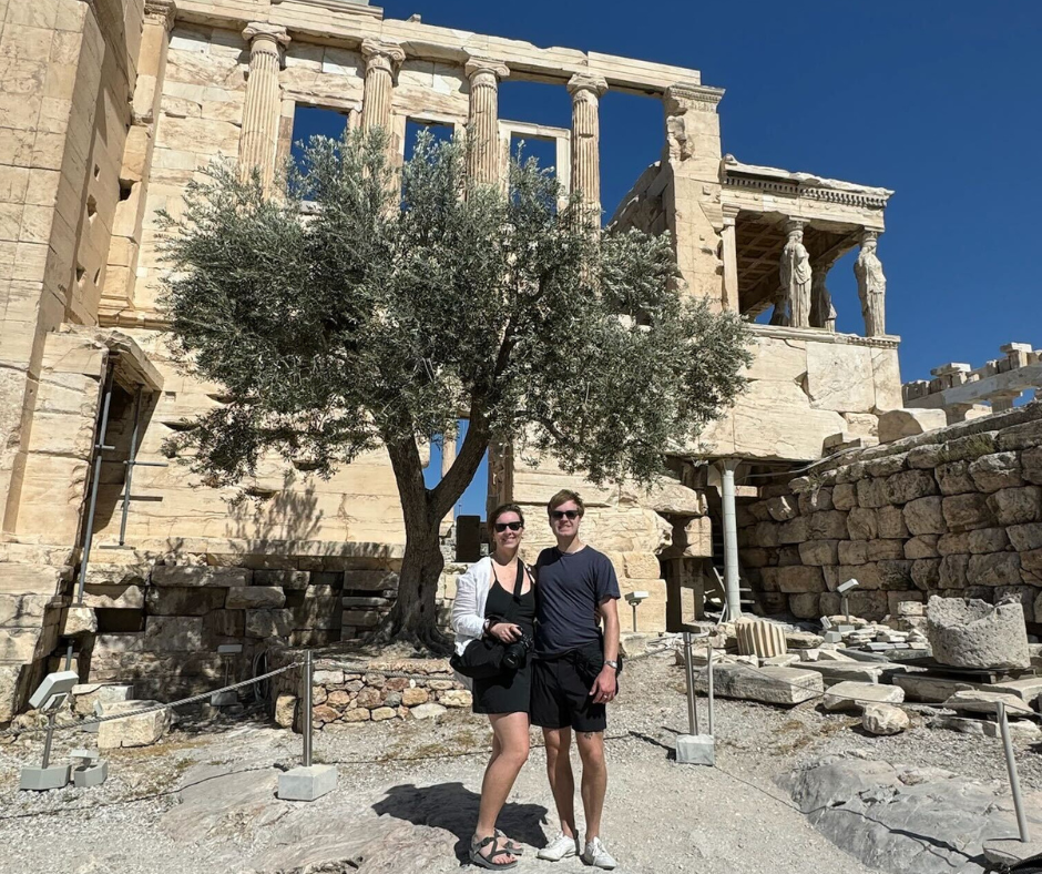 Couple stands in front of olive tree with ancient marble temple in background.