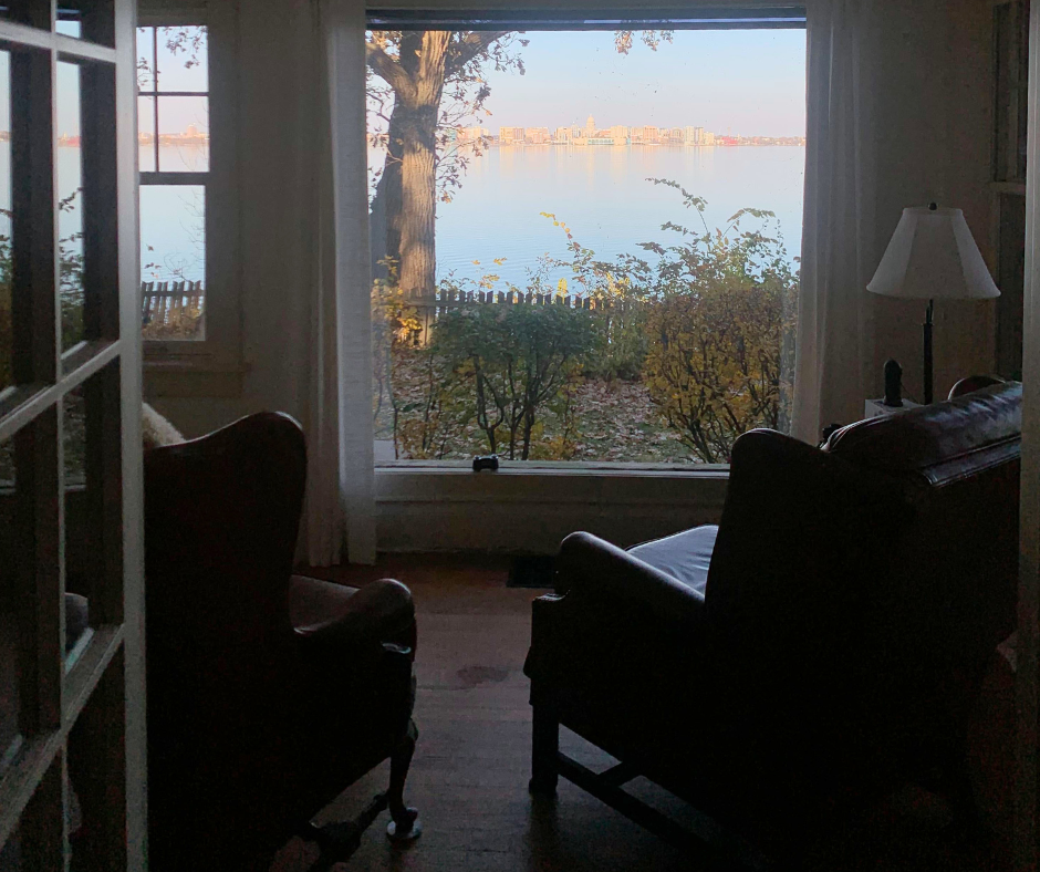 View of lake out of window on sunny day. City in background. Leather chairs and wood floor look out window over the water.