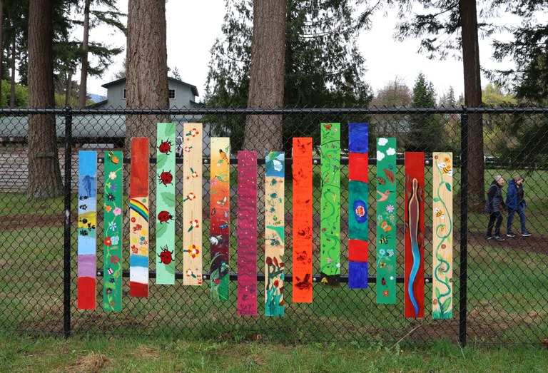 Hand painted wood panels detailing local pollinators like bees cover black chain link fence. Grassy, gravel trail with tall pine trees in background. Two hikers walking to the left hand side.