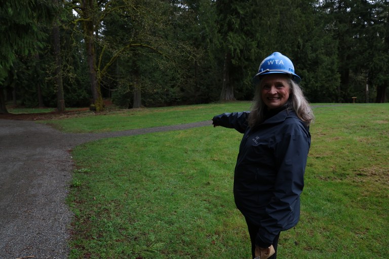 Women wearing blue hard hat points to gravel trail amongst grass. Evergreens in background.