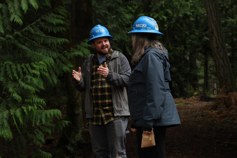 Two people in hard hats talk with evergreens in background. 
