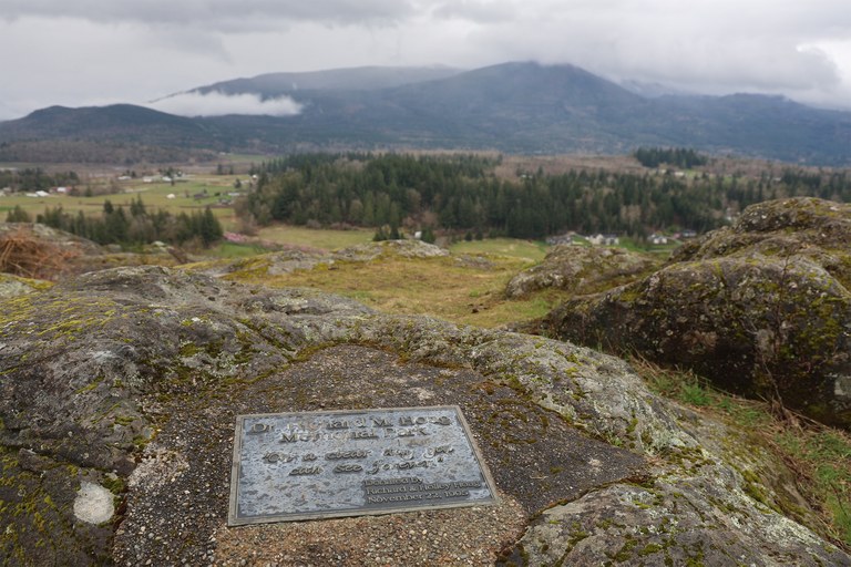 A plaque on a rock over a view of a valley with hills and low clouds beyond. 