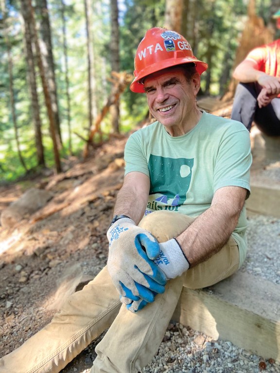 A volunteer in an orange hard hat sits on a step on trail and smiles at the camera. 