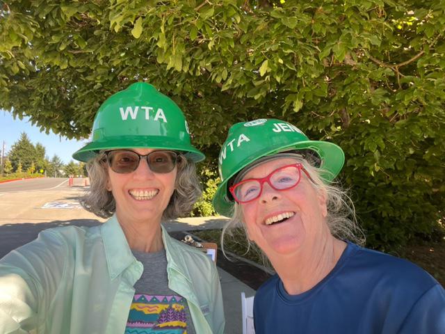 Two women pose outside with a tree in the background. They are wearing green WTA heard hats.