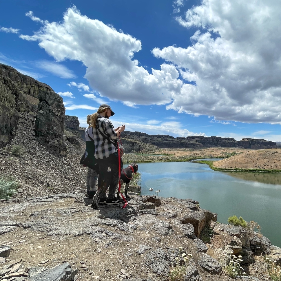 Two adults and a leashed dog standing at the edge of the rocks taking a photo of the water at Ancient Lakes. Photo by Leap.