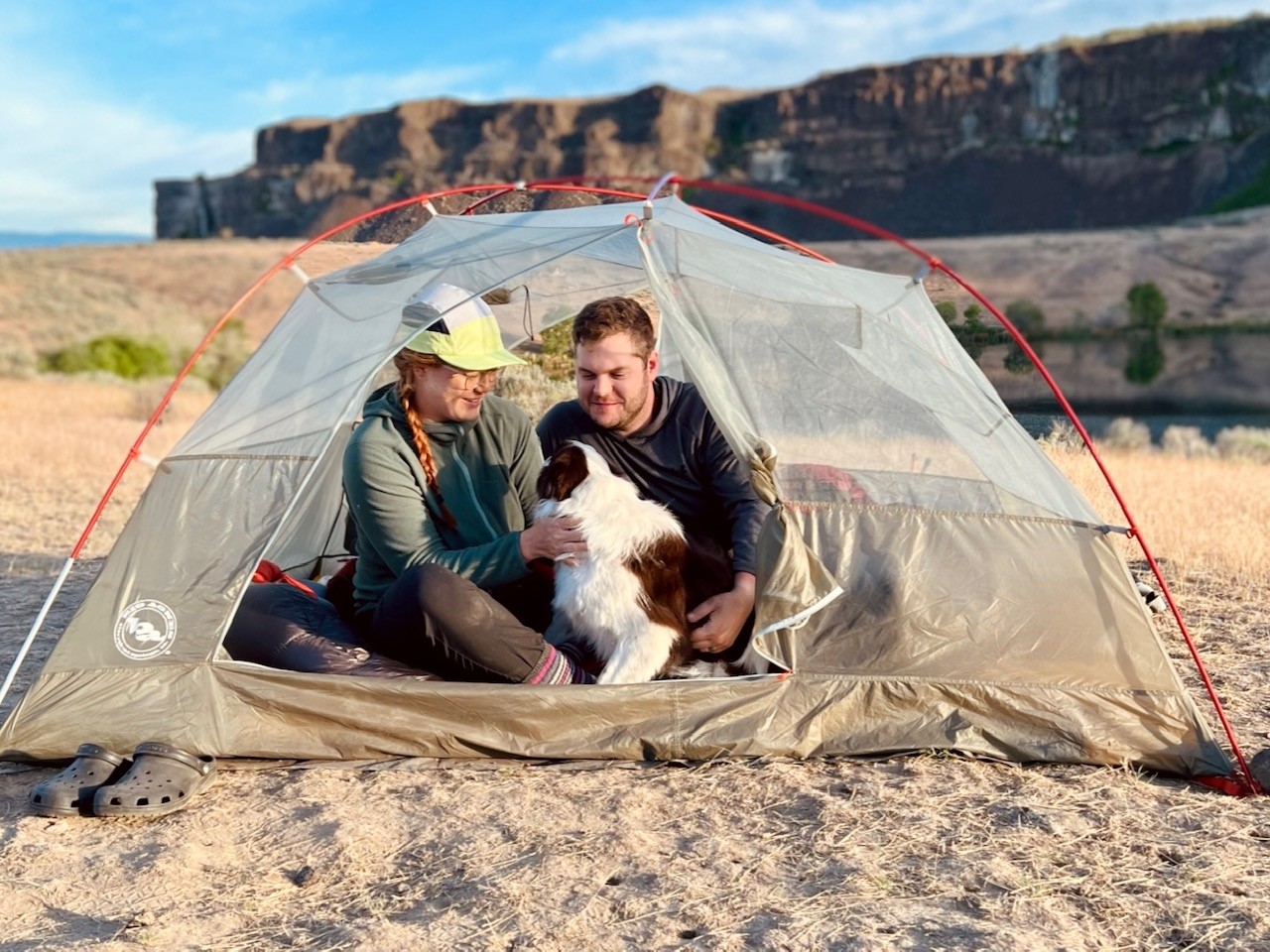 Two people sit smiling with a dog in a tent with a desert bluff behind them