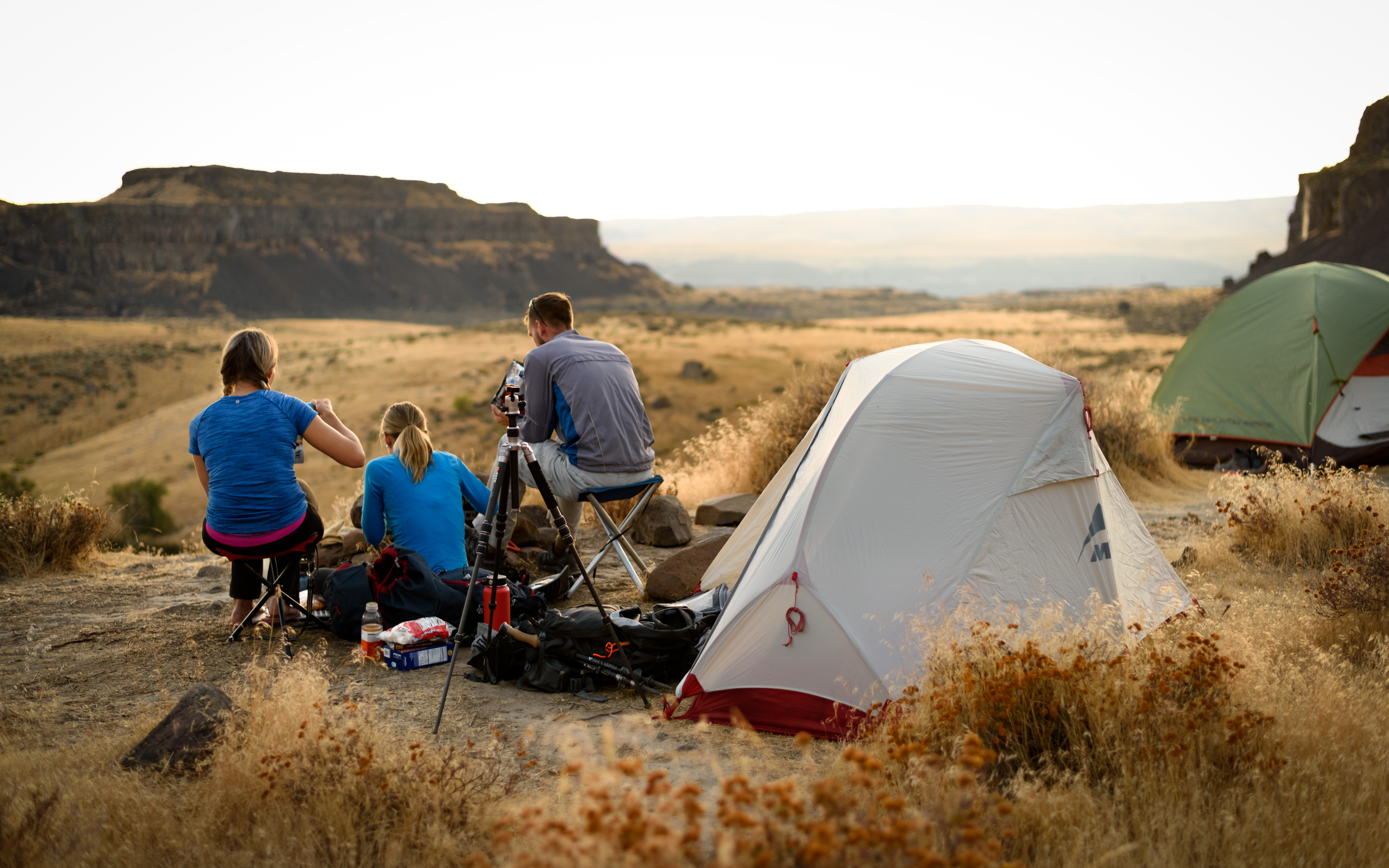Three people sit next to a tent in a desert landscape with plateaus in the distance