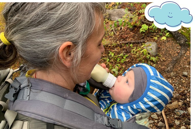 A hiker feeds a bottle to a baby in a front carrier. 