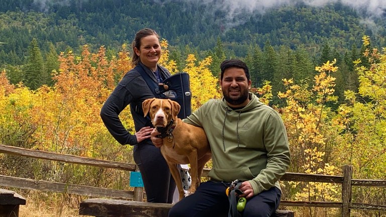 A hiker stands behind a bench with a baby in a baby carrier on her chest. Another hiker sits on the bench, with his arm around a large orange dog. Fall colors and evergreen are visible in the background. 