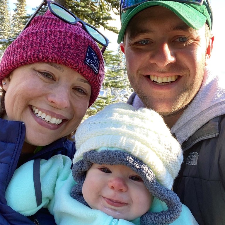 Three hikers, two adults and a baby, all smile at the camera for a selfie. Everyone is wearing hats and the baby is wearing a cozy fleece outer layer. 