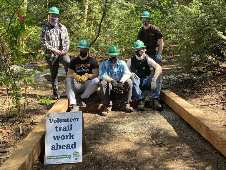 Five volunteers, wearing WTA green hard hats, sit and stand amid a partially finished structure to lift the trail out of a wet area. 