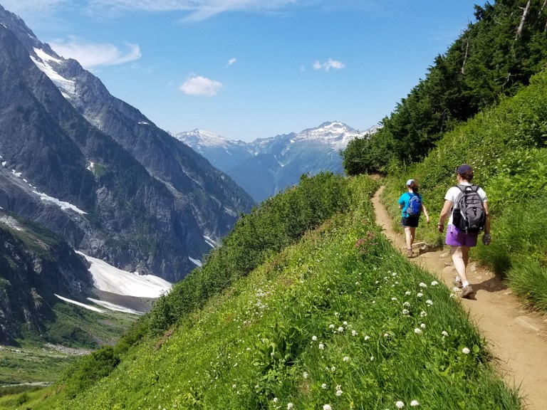 Two hikers on a dirt trail surrounded by grass, with mountains in the background. Photo by Bradley Mitchell.