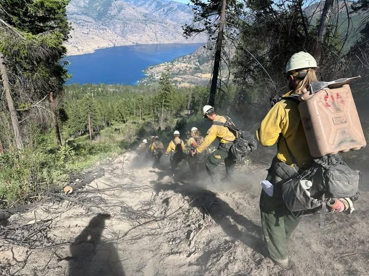 Line of firefighters building a fireline down a steep slope to a lake.