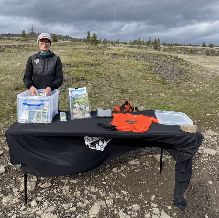 Ambassador at Catherine Creek Ambassador poses behind an outreach table at the Catherine Creek Trailhead. The tablecloth is blowing in the wind.