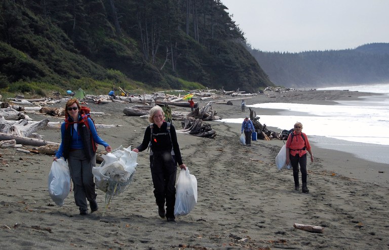 Several women walk toward the camera on a beach carrying full trash bags and smiling. 