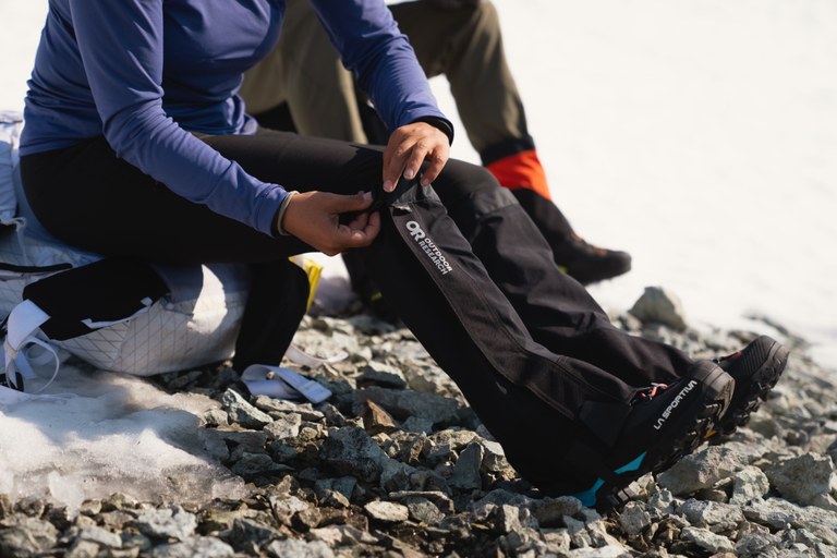 A hiker puts on OR gaiters will sitting at the edge of a patch of snow.