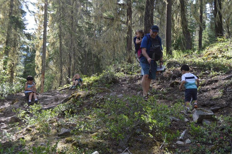 Cooper River. Photo by Ups N Downs. A man wearing a child in a backpack leans down toward another child walking toward him. Two kids are in the background. They are all in a forest area.