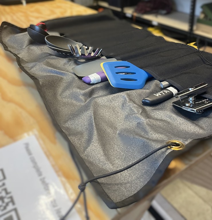 A black rectangular piece of fabric has sewn pockets which each contain a variety of utensils