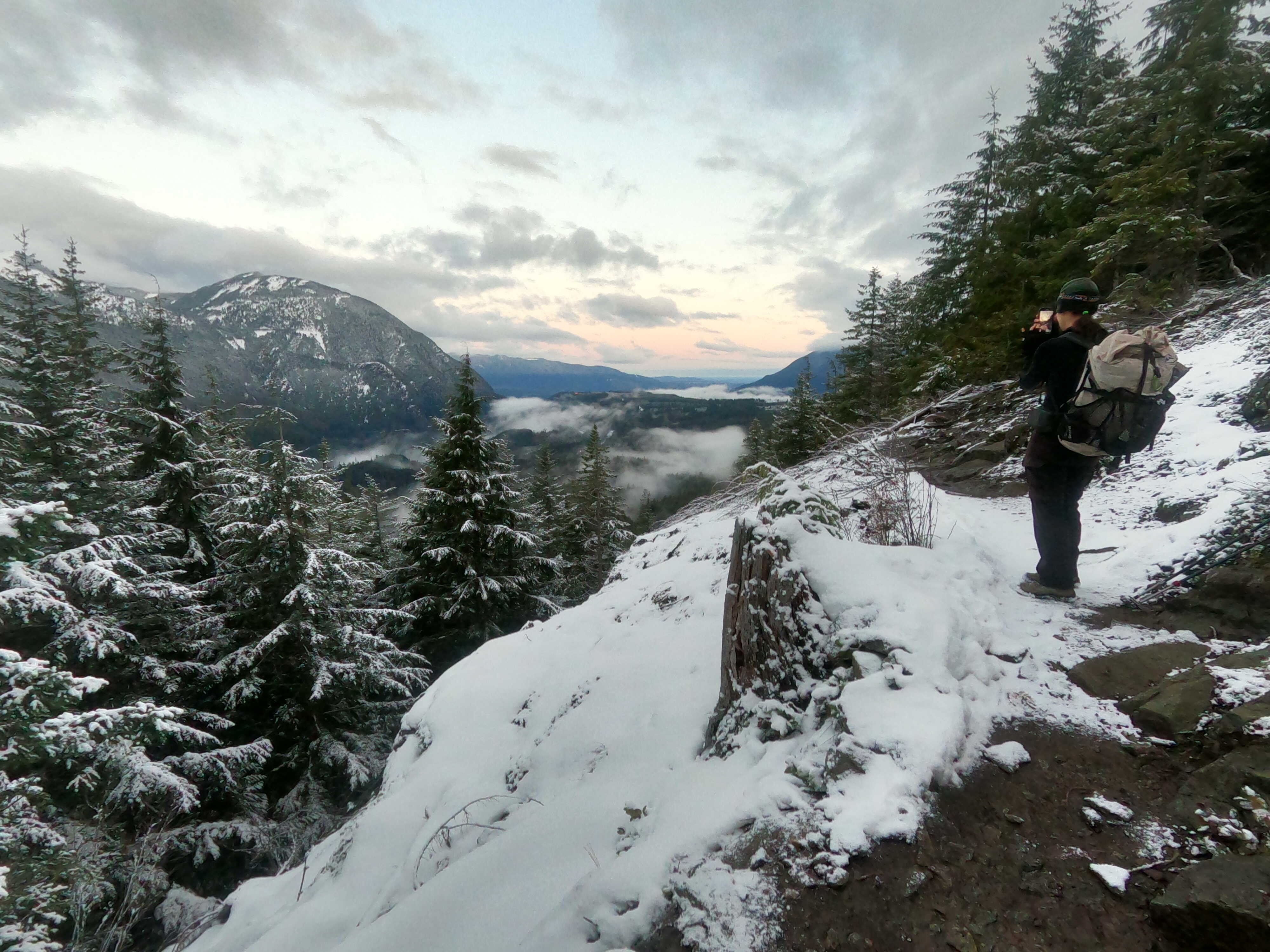 Hiker stands on a snowy trail and takes a photo of the surrounding trees, rolling mountains, pastel sky