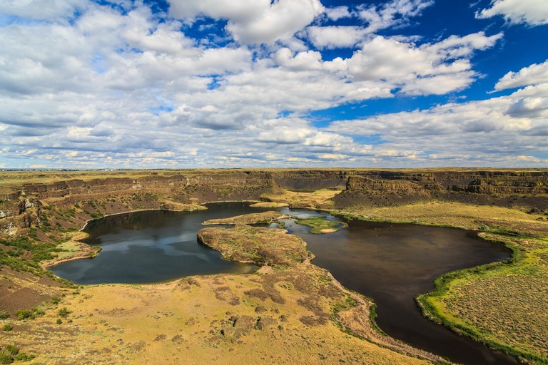 Huge cliffs, which were once waterfalls, with lakes and a desert landscape in front and a blue sky with white clouds behind.